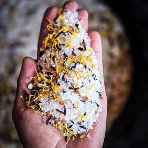 May include: A hand holding a mixture of white bath salts with dried yellow and red flower petals.