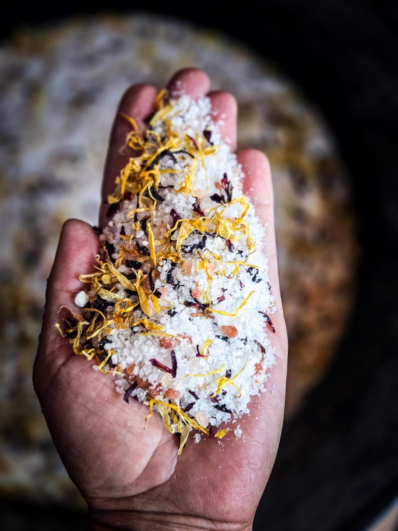 May include: A hand holding a mixture of white bath salts with dried yellow and red flower petals.