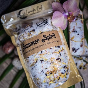 May include: A clear plastic bag filled with a white bath salt blend with dried flowers and herbs. The bag has a brown paper label that says "Summer Spirit" and lists the ingredients: Dead Sea Salt, Epsom Salt, Coconut, Hibiscus, Mango.