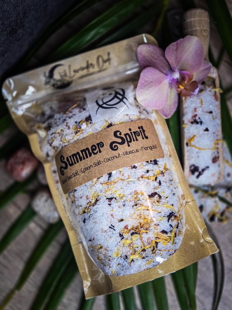 May include: A clear plastic bag filled with a white bath salt blend with dried flowers and herbs. The bag has a brown paper label that says "Summer Spirit" and lists the ingredients: Dead Sea Salt, Epsom Salt, Coconut, Hibiscus, Mango.