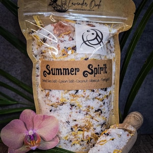 May include: A brown paper bag filled with a white bath salt blend called "Summer Spirit". The bath salt blend contains Dead Sea Salt, Epsom Salt, Coconut, Hibiscus, and Marigold. The bag is sitting on a wooden surface with a pink orchid and a wooden scoop.