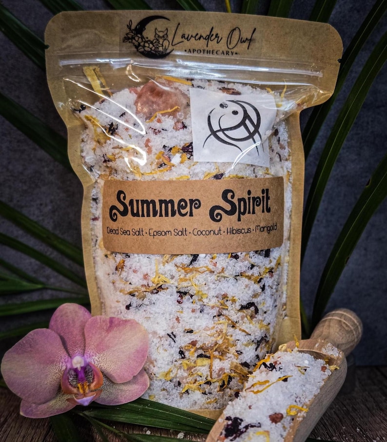 May include: A brown paper bag filled with a white bath salt blend called "Summer Spirit". The bath salt blend contains Dead Sea Salt, Epsom Salt, Coconut, Hibiscus, and Marigold. The bag is sitting on a wooden surface with a pink orchid and a wooden scoop.