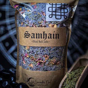 May include: A brown paper bag of Samhain Ritual Bath Salts, with a clear window revealing the contents of dried flowers and herbs. A wooden scoop holds more herbs. Black stones are in the foreground.