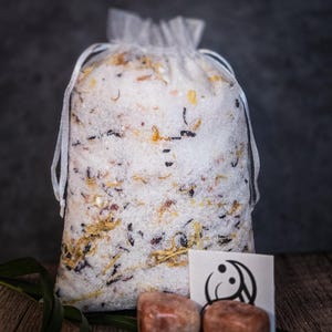 May include: A white mesh bag filled with bath salts and dried flowers. The bag is sitting on a wooden surface with two small, orange-brown stones and a white card with a black yin-yang symbol.