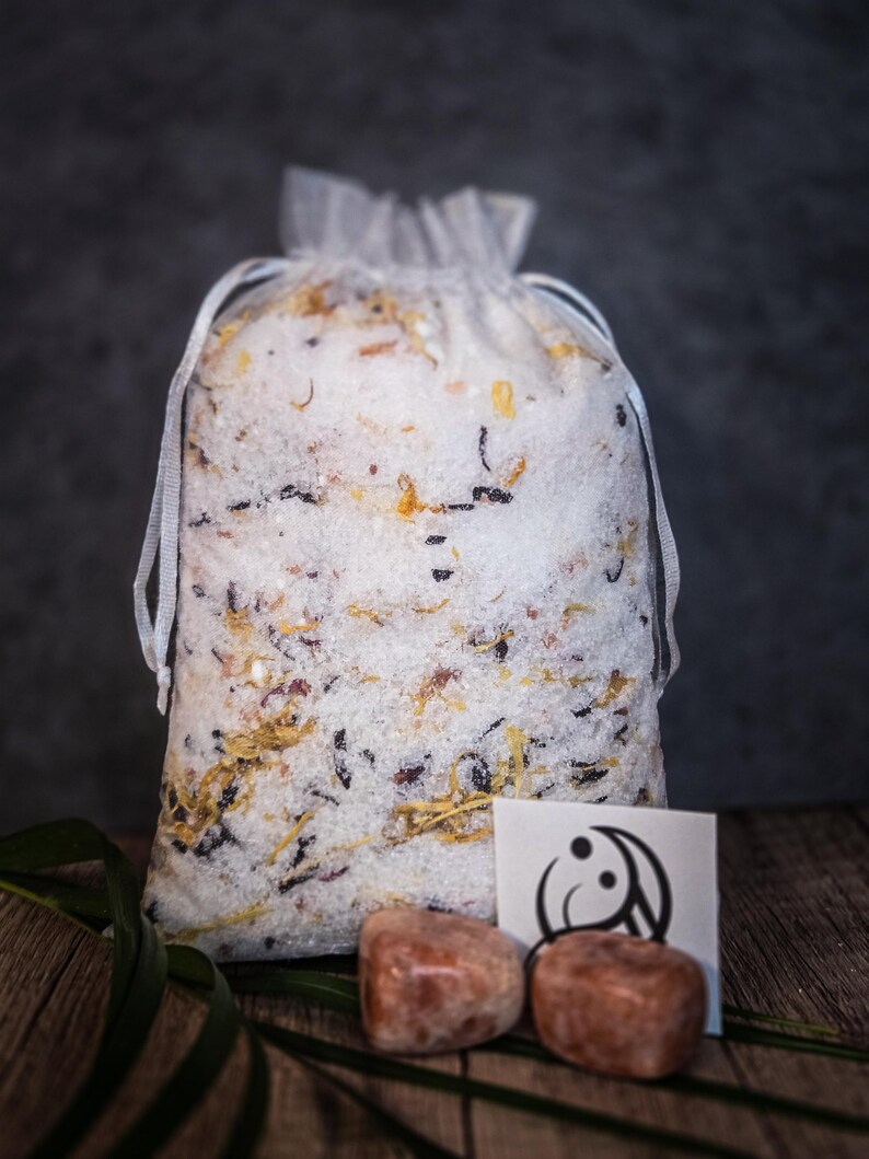 May include: A white mesh bag filled with bath salts and dried flowers. The bag is sitting on a wooden surface with two small, orange-brown stones and a white card with a black yin-yang symbol.