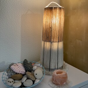 May include: A white and brown woven lampshade with a white fringe base sits on a white table next to a pink Himalayan salt lamp with a white fringe base. A blue wire basket filled with smooth stones and a pink crocheted heart sits on the table.