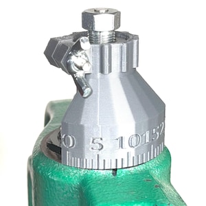 May include: A close-up of a gray metal tool with a numbered dial that reads "0 5 10 15 2". The tool is mounted on a green base.