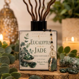 May include: A square glass reed diffuser bottle with a label reading LYNNMARIE EUCALYPTUS JADE REED DIFFUSER 2 OZ (59 mL). The label features a green eucalyptus illustration and a bowl of jade stones. A gold chain with a letter L charm and a raw jade crystal hangs from the black cap, which holds several wavy brown reeds.
