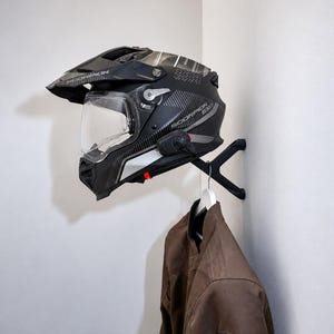 Peut inclure: Un casque de moto noir Scorpion EXO avec une visière transparente et un dispositif de communication fixé sur un support mural noir. Sous le casque, une veste marron est suspendue à un cintre blanc sur la même structure de support murale.