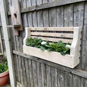 May include: A wooden planter box with white flowers, mounted on a wooden fence. The planter box has a slatted design and is made from reclaimed wood.
