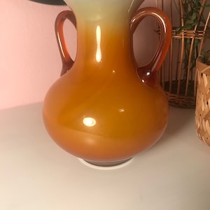 May include: A large, amber-colored glass vase with two handles. The vase has a gradient of color, starting with a light cream at the top and fading to a deep amber at the bottom.