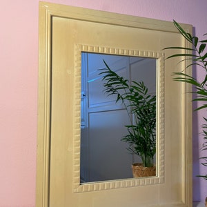 May include: A rectangular wall mirror featuring a wide, light cream-colored wooden frame. The inner border of the frame is decorated with a repeating square-patterned relief detail. The reflective glass surface shows a reflection of a green houseplant.