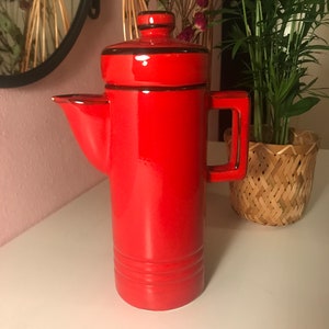 May include: A red ceramic teapot with a black lid and handle. The teapot has a cylindrical body with a rounded bottom and a spout on the side. The teapot is sitting on a white surface.