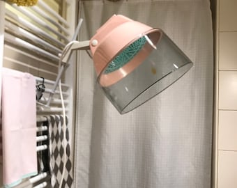 Puede incluir: Un secador de pelo con capucha de plástico rosa y transparente con un brazo blanco unido a un toallero de pared blanco. La capucha está inclinada hacia abajo y el brazo está extendido.