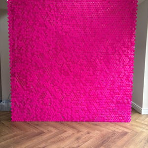 May include: A large, bright pink wall decoration made up of many small, hexagonal elements. The wall has a uniform colour, creating a textured, striking display. The background shows a wooden floor and white walls.