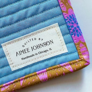Könnte beinhalten: Ein blauer gesteppter Stoff mit einem weißen Etikett, auf dem "Quilted by Aimee Johnson Handmade in Chicago, IL" steht. Der Stoff hat einen rosa-goldenen Blumendruck am Rand.