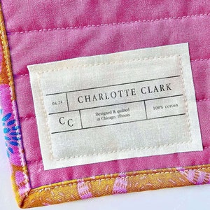 Puede incluir: Una etiqueta blanca con el texto "CHARLOTTE CLARK" en letras negras. La etiqueta también incluye el texto "Designed & quilted in Chicago, Illinois" y "100% algodón". La etiqueta está unida a una tela rosa y naranja.