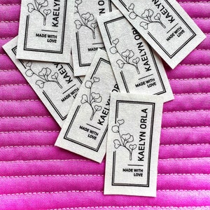 May include: Close-up of several white fabric labels with black text and a simple line drawing of a plant. The text on the labels reads "Kaelyn Orla" and "Made with Love".