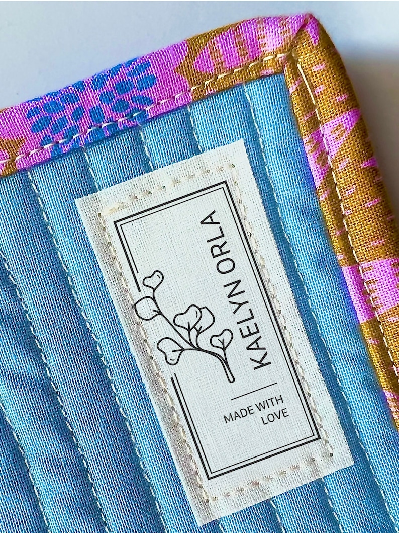 May include: A white fabric label with black text that reads "Made with Love" and "Kaelyn Orla" with a small floral design. The label is sewn onto a blue fabric with a pink and gold patterned border.