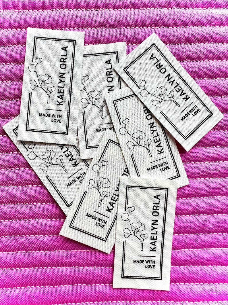May include: Close-up of several white fabric labels with black text and a simple line drawing of a plant. The text on the labels reads "Kaelyn Orla" and "Made with Love".