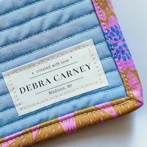 May include: A blue quilted fabric with a white label that reads "DEBRA CARNEY, Madison, WI" and the text "created with love +" . The fabric has a pink and gold floral pattern on the edge.