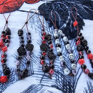 May include: Several beaded necklaces with a gothic theme. The necklaces feature black skull beads, red rose beads, and black rose beads. Some necklaces have white skull beads and purple beads.