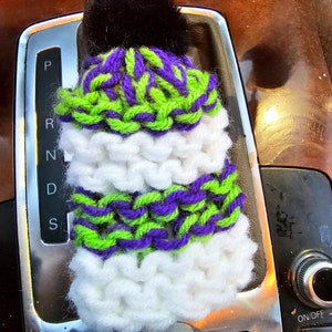May include: A white, green, and purple knitted car gear shift knob cover with a black pom pom on top.