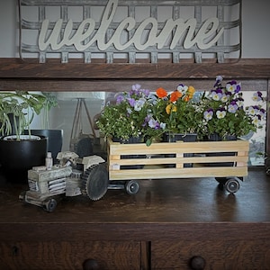 May include: A wooden wagon with wheels, carrying a planter box filled with purple, white, and orange pansies. The wagon is on a dark wood surface with a toy tractor in front of it. A sign that says "welcome" is hanging on the wall above the wagon.