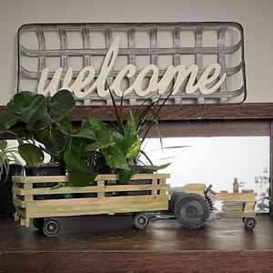 May include: A wooden toy tractor with a green and brown paint finish, carrying three potted plants. The tractor has black wheels and is sitting on a wooden shelf with a mirror behind it. A metal sign with the word "welcome" is hanging above the shelf.