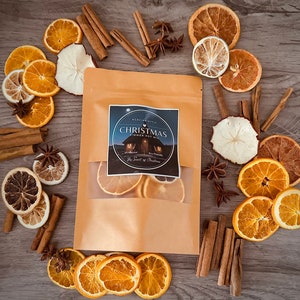 May include: A kraft paper bag of Christmas simmer pot mix with dried orange slices, cinnamon sticks, star anise, and apple slices. The bag has a label with the text "Christmas Simmer Pot Mix" and "The Scent of Christmas". The items are arranged on a wooden surface.