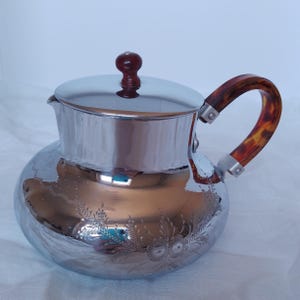 May include: A polished chrome teapot featuring a rounded body with an etched floral pattern, a matching lid with a small brown knob, and a curved handle with a brown and orange tortoiseshell-style pattern attached with silver-toned metal hardware.