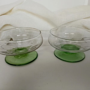 Può includere: Due coppette da dessert in vetro trasparente con base verde. Le coppette hanno una forma arrotondata e poggiano su una base verde circolare. Lo sfondo presenta un tessuto bianco strutturato.