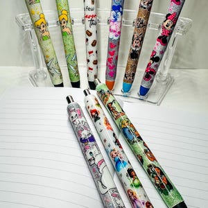 May include: A collection of decorative pens with various designs, including cartoon characters and patterns. The pens are displayed on a clear stand and a notebook. The pens feature glitter accents and black ink tips.