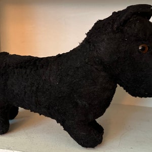 May include: Black plush toy dog with brown button eyes. The dog is standing on a white surface.