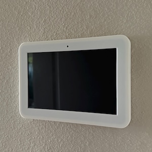 May include: A white rectangular smart home control panel with a black screen is mounted on a textured beige wall. The panel has rounded corners and a small camera lens at the top.