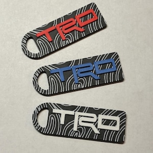 May include: Three TRD keychains with a black background and a white swirl pattern. The TRD logo is in red, blue, and white. Each keychain has a hole at one end.