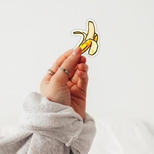 Puede incluir: Una mano sosteniendo una pegatina de un plátano pelado. La pegatina del plátano es amarilla y blanca con contornos negros. La mano tiene dos anillos en los dedos y lleva una sudadera gris.