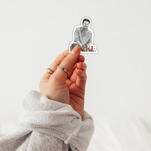 May include: A sticker featuring a black and white photo of a man with brown hair and a beard. The man is wearing a light blue shirt and jeans. The sticker has the word "JAKE" printed in brown letters below the photo.