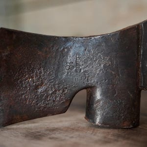 Puede incluir: Una cabeza de hacha vintage oxidada con un mango de madera. La cabeza del hacha tiene un pequeño agujero en el lateral y el texto "81 GUTTE" está estampado en el lateral.