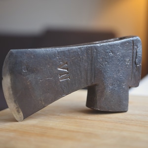 Peut inclure: Une hache vintage, patinée, avec une tête en métal et un manche en bois. La tête de la hache porte une petite marque rectangulaire avec les lettres "VII" inscrites dessus.