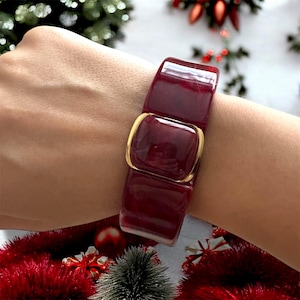 May include: A burgundy bracelet with a gold-colored clasp. The bracelet is made of rectangular, glossy pieces. The clasp is square and connects the two sections of the bracelet. The bracelet is worn on a wrist.