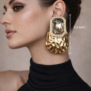 May include: Gold-coloured statement earrings with a large, rectangular smoky gemstone centre, surrounded by smaller clear stones. The earrings are approximately 6.1 cm in length and feature a textured, hammered metal design. The brand name "MAISON NURÉE" is at the top.