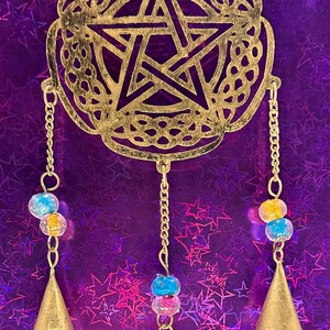 May include: A gold-colored metal wind chime with a pentagram design. The chime has three chains with colorful beads and small bell-shaped pendants. The background is a purple, star-patterned surface.