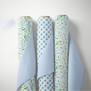 May include: Three rolls of white fabric with a blue and green floral pattern. The fabric is rolled and partially unrolled, revealing the pattern. The fabric is displayed against a white background.