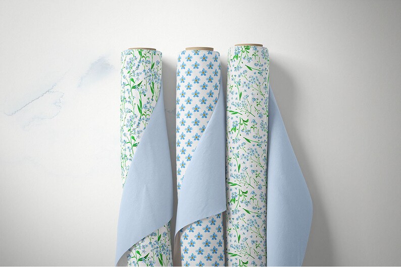 May include: Three rolls of white fabric with a blue and green floral pattern. The fabric is rolled and partially unrolled, revealing the pattern. The fabric is displayed against a white background.