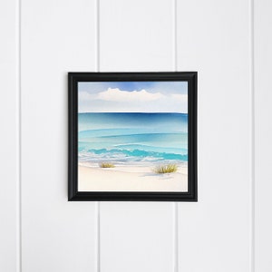 Puede incluir: Una pintura de acuarela enmarcada de una escena de playa. La obra de arte representa un oc&eacute;ano sereno con tonos de azul y turquesa, una playa arenosa y un cielo nublado. La pintura est&aacute; encerrada en un marco negro y montada en una pared blanca.