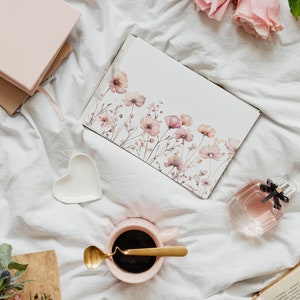 K&ouml;nnte beinhalten: Eine rosa Kaffeetasse mit einem goldenen L&ouml;ffel auf einem wei&szlig;en Bettlaken. Eine Aquarellkarte mit rosa Blumen liegt auf dem Bettlaken. Eine durchsichtige Glasflasche mit Parf&uuml;m liegt auf dem Bettlaken. Ein rosa Notizbuch liegt auf dem Bettlaken.