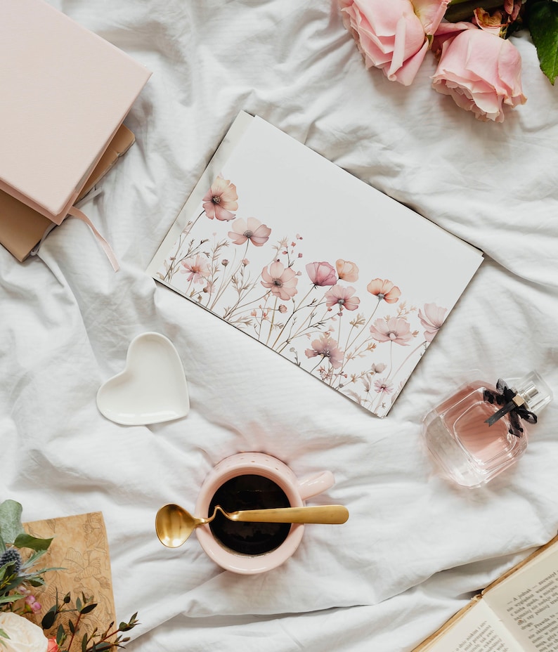 K&ouml;nnte beinhalten: Eine rosa Kaffeetasse mit einem goldenen L&ouml;ffel auf einem wei&szlig;en Bettlaken. Eine Aquarellkarte mit rosa Blumen liegt auf dem Bettlaken. Eine durchsichtige Glasflasche mit Parf&uuml;m liegt auf dem Bettlaken. Ein rosa Notizbuch liegt auf dem Bettlaken.