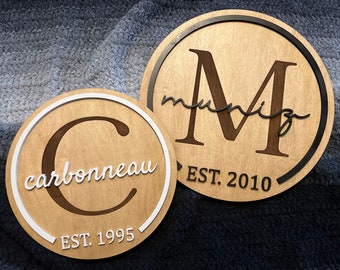 May include: Two wooden signs with black and white trim. The first sign has a large "C" with the text "carbonneau" and "EST. 1995". The second sign has a large "M" with the text "muniz" and "EST. 2010".