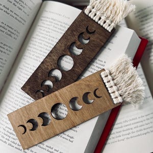 May include: Two wooden bookmarks with a moon phase design. The bookmarks are rectangular with cut-out crescent moon shapes and a cream-colored tassel at the top. One is light brown, the other is dark brown, and both are resting on an open book.